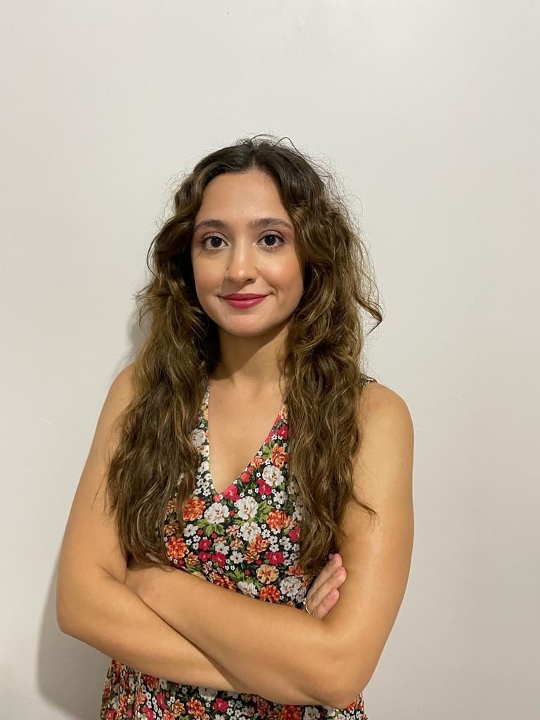 Person with long, wavy hair wearing a floral sleeveless dress, standing with arms crossed.